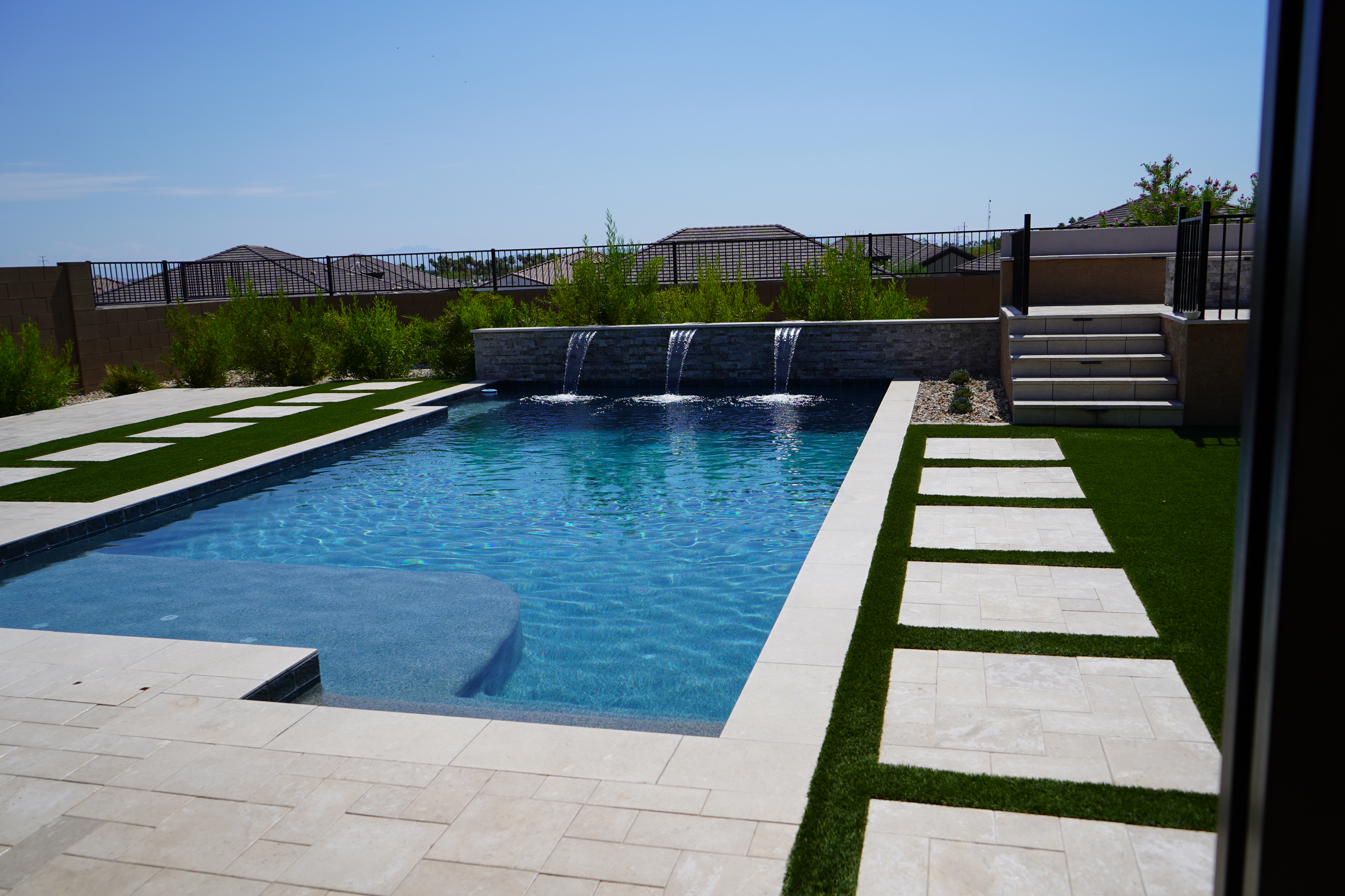 Modern backyard pool with raised stone water wall, waterfalls, turf paver path, and desert mountain view
