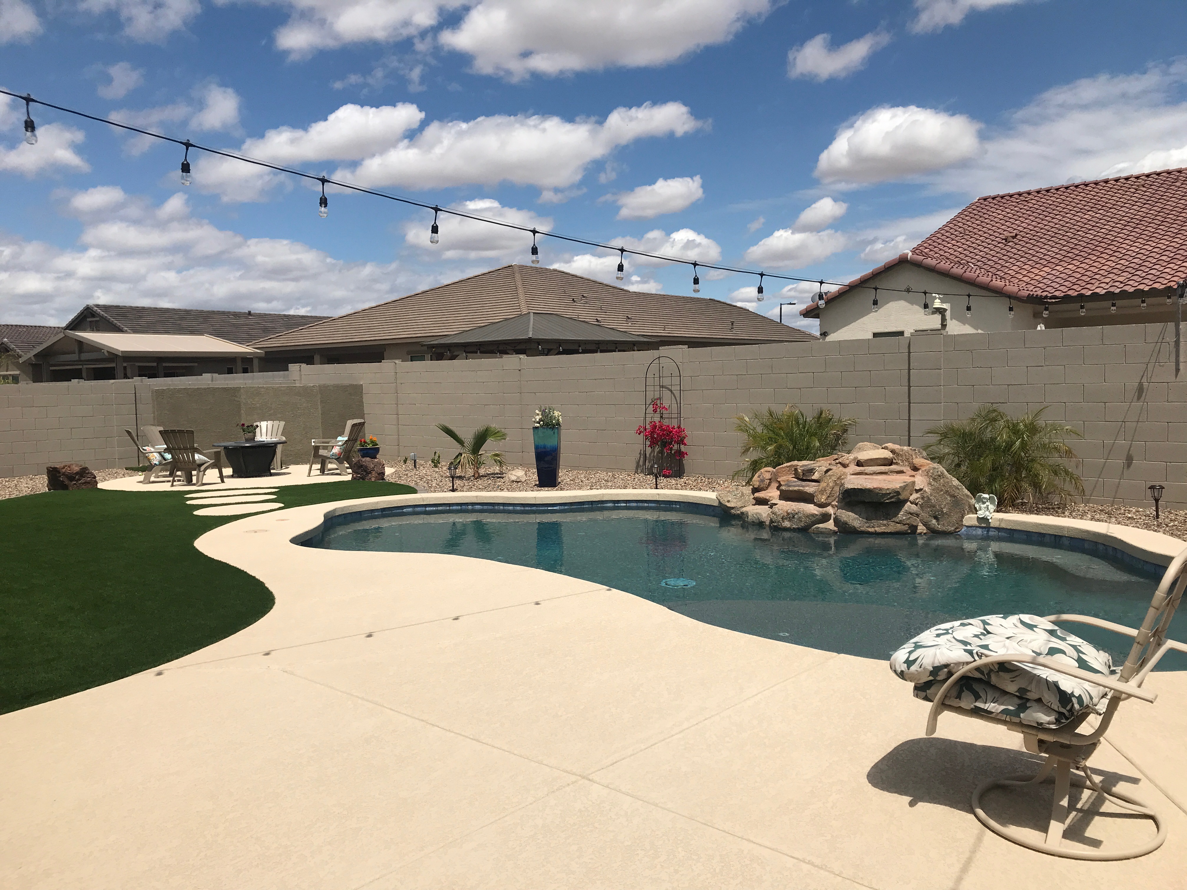 Curved backyard pool with boulder waterfall, patio lounge furniture, grass, and string lights