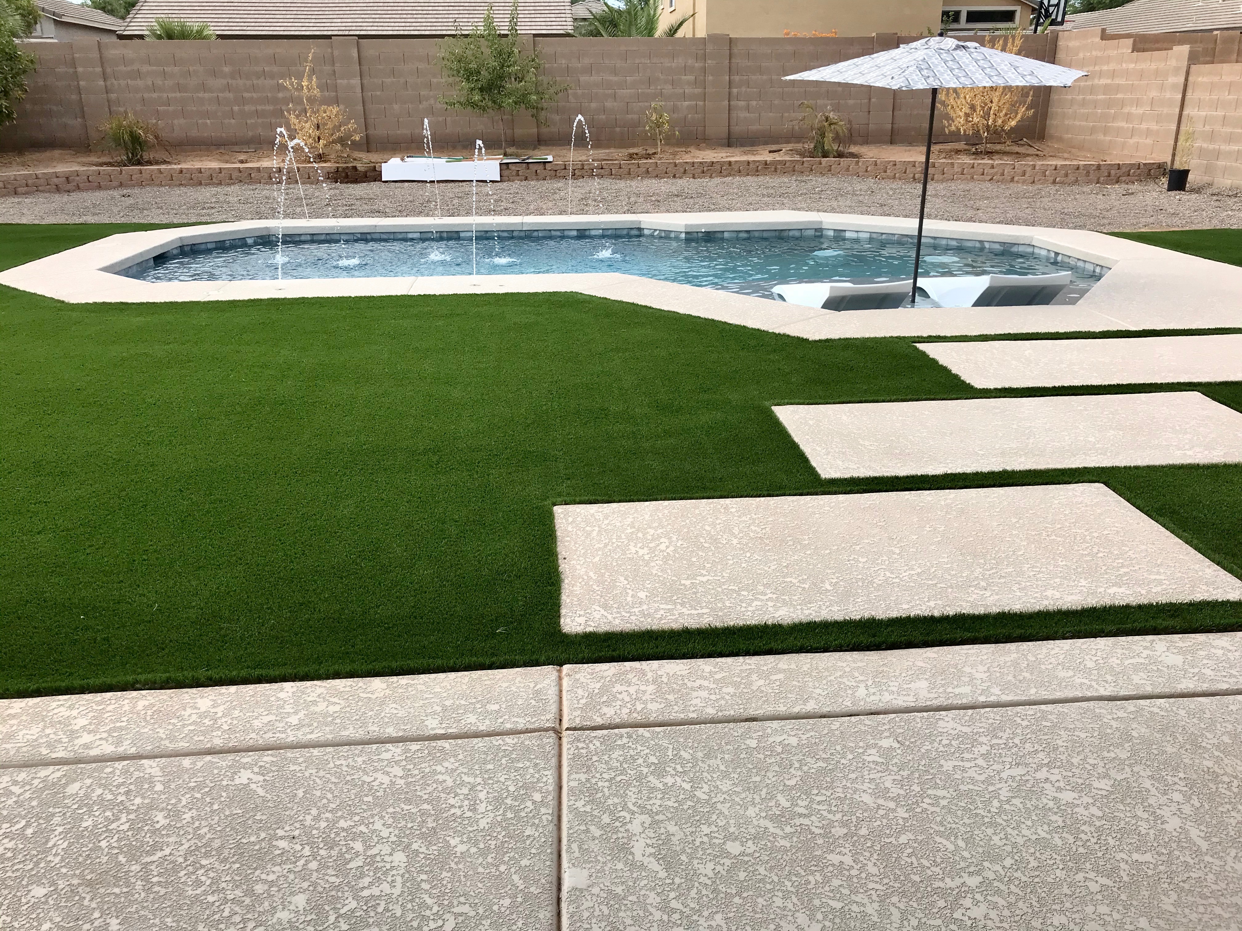 Wide backyard pool with bubbler jets, umbrella, submerged lounge chairs, turf, and concrete stepping pads