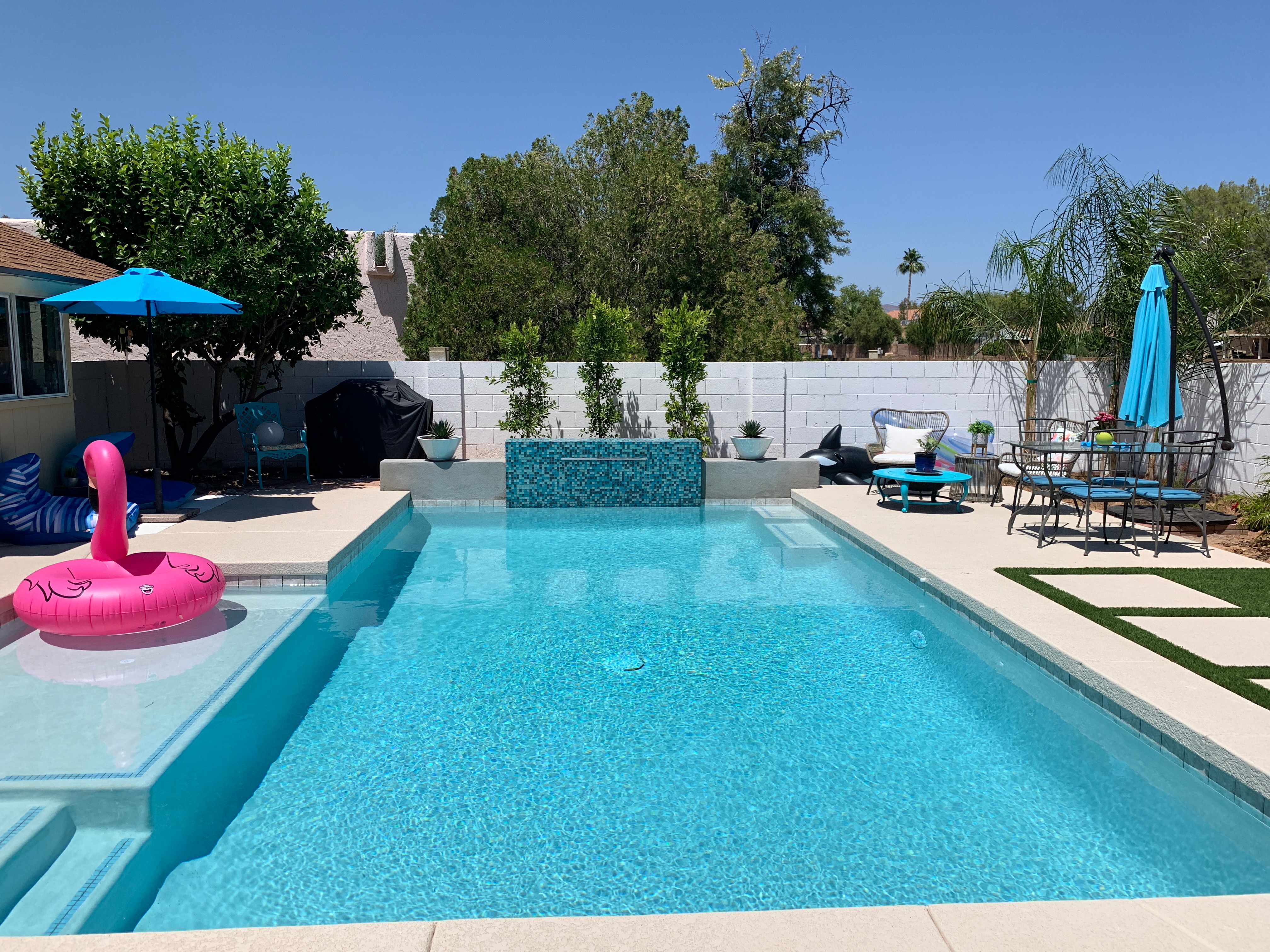 Bright backyard pool with turquoise tile wall, shallow ledge, patio furniture, umbrellas, and lounge float